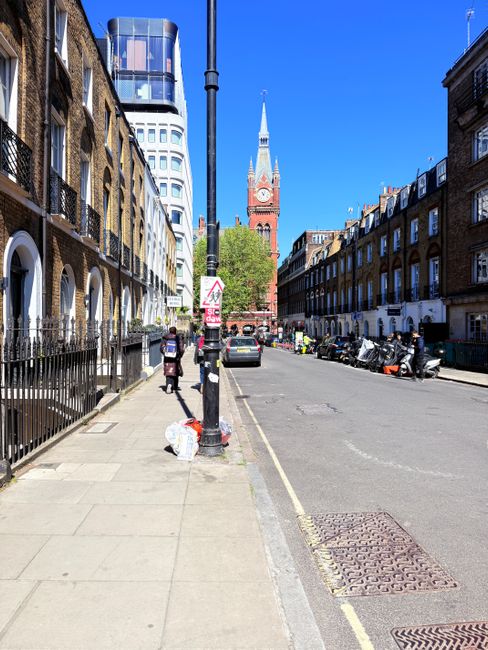 A narrow cobbled street in Bermondsey with brick buildings on both sides and black bollards lining the pavement. Several decorative iron pedestrian bridges span across the alleyway, connecting the bui