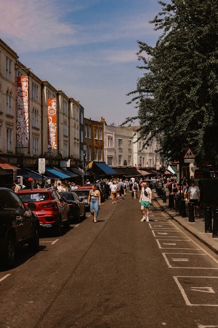 A busy street scene on Bermondsey Street during daytime, with several multi-storey buildings on the left side featuring signage and shopfronts. The pavement is occupied by pedestrians, some carrying boxes or bags, while others walk or wait near parked vehicles. Multiple cars are lined up along the curb, with a red car visible in the foreground. On the right side, there is a designated parking area with marked spaces and black bollards, separated from the pedestrian walkway by a row of short black posts. Large leafy trees shade parts of the street, and the sky is partly cloudy with patches of blue. The scene suggests a bustling environment suitable for house or business relocations, with ongoing loading or packing activity potentially linked to house removals or furniture transport, as supported by the presence of cardboard boxes, soft packaging materials, and the general movement of items within the area. Man with Van Bermondsey is experienced in handling such home relocation logistics, ensuring professional packing and moving services in environments like this.