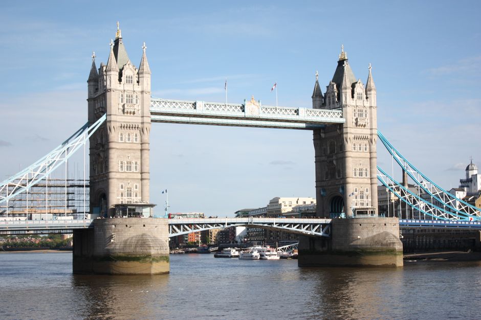 The image shows the Tower Bridge in London with its two towers and suspension spans, set against a partly cloudy sky. The bridge is positioned over the River Thames, with the water reflecting the stru